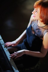 Fototapeta na wymiar Beautiful cheerful girl, illuminated by a beam of bright light, sits at the piano and smiles genuinely on a dark background closeup