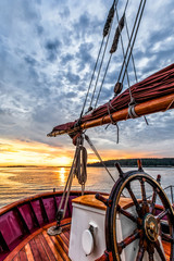 Obraz premium Sunrise at sea on a tall ship classic sailboat. Close up of the wheel, boom and stern against a dramatic sky, clouds and the gold light of dawn reflected in the water.