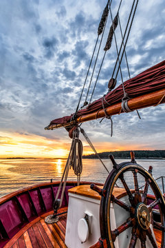 Sunrise at sea on a tall ship classic sailboat. Close up of the wheel, boom and stern against a dramatic sky, clouds and the gold light of dawn reflected in the water.