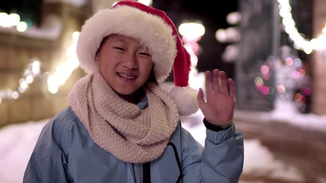Happy Little Girl, Wearing Santa Hat, Waves In Front Of Festive Winter Holiday Scene (Slow Motion) 
