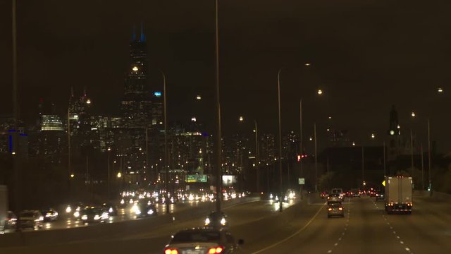POV: Driving on scenic heavy traffic interstate highway towards the iconic Loop, central business district illuminated with colorful lights at night. People on road trip traveling to downtown Chicago