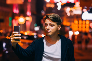 Young man take selfie on smartphone while walking in night city streets. Smiling guy on blurred lights background