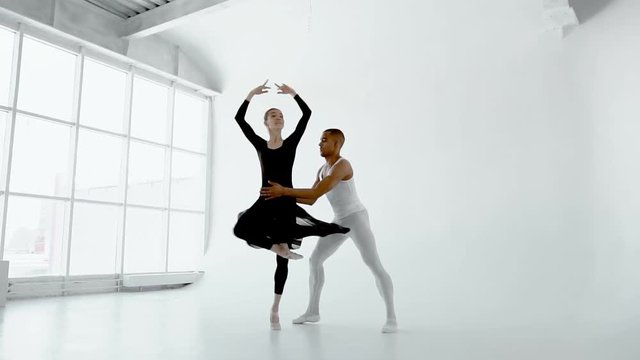 Young 20s ballet dancer gently holds the attractive brunette ballerina during the dance in white dance hall with big window slow motion 120fps.