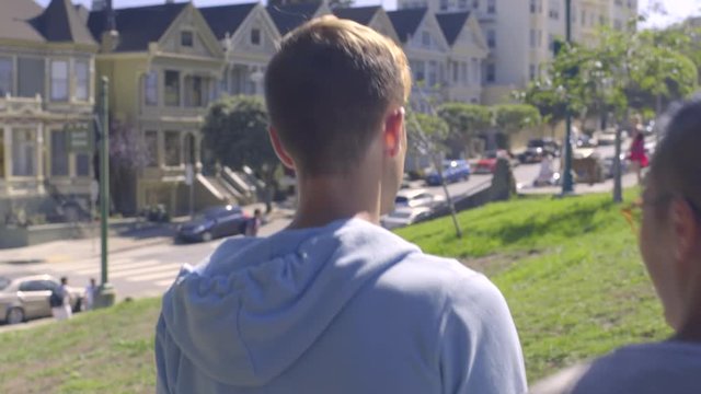 Gay Couple Walk Together In Alamo Square, San Francisco, Man Points To Historic Painted Ladies Across Street 