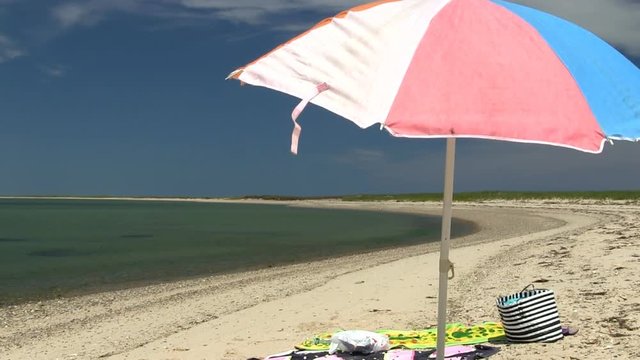 A colorful beach umbrella against blue sky moves in light breeze.  A secluded stretch of beach coastline at south monomoy island Cape Cod