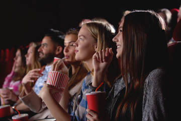 Group of female friends laughing and smiling watching a film together at the movie theatre snacks popcorn entertainment people lifestyle leisure friendship concept. - obrazy, fototapety, plakaty