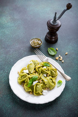 Homemade Italian tortelloni with green pesto, goat cheese and pine nuts on white plate on green stone background. Healthy food concept. Copy space. High angle view.