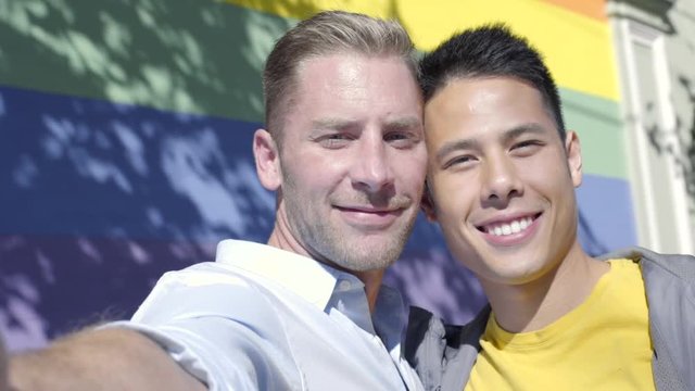 Attractive Gay Couple Kiss On Cheek And Pose For Selfie In Front Of Gay Pride Rainbow Wall