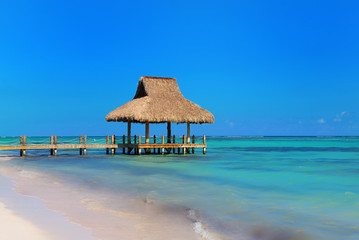 Tropical white sandy beach. Palm leaf roofed wooden pier with gazebo on the beach. Punta Cana, Dominican Republic