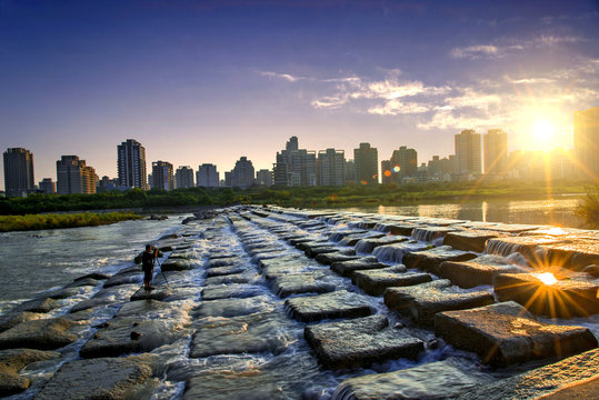 White water cascades over square block with city skyline in the background, A slow exposure shows the motion of the flowing water.
