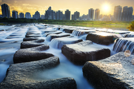 White Water Cascades Over Square Block With City Skyline In The Background, A Slow Exposure Shows The Motion Of The Flowing Water.