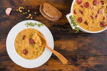 Hearty and healthy split pea and smoked sausage soup with green thyme in a white plate and casserole on the wooden table, top view.