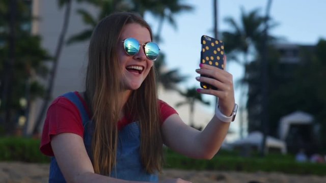 Happy Teen Girl Poses For Fun Selfies On The Beach, Resort With Palm Trees In Background, She Looks At The Photo After