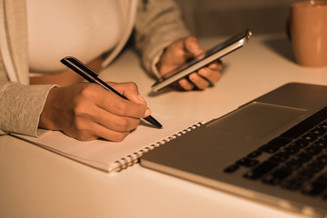 cropped shot of young woman writing at notebook while holding smartphone at home