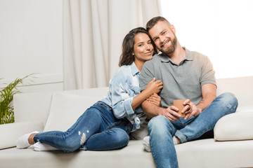 young happy couple looking at camera while hugging on sofa at home