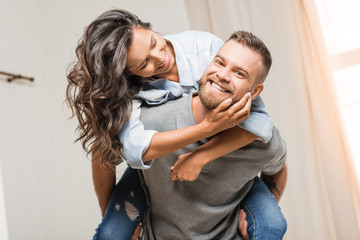 portrait of happy man looking at camera while piggybacking with woman at home