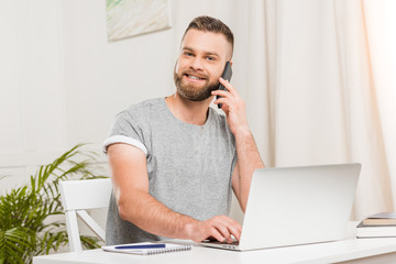 portrait of smiling man talking on smartphone while typing on laptop