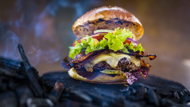 Tasty smoked grilled and glazed beef burger with lettuce, cheese and bacon served with french fries on wooden table with copyspace, smoke mesquite timber wood in background.