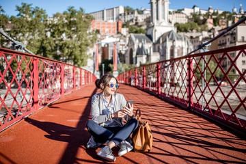 Young stylish busineswoman sitting with phone and bag on the red footbridge in Lyon, France