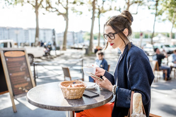 Young woman having a breakfast with coffee and croissant sitting outdoors at the french cafe in Lyon city - obrazy, fototapety, plakaty