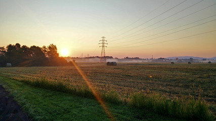 Electrical poles and bales of straw on farmland in a foggy summer morning with sunrise in the background