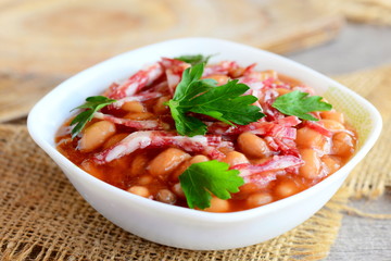 Smoked sausages with white beans in tomato sauce. Simple white bean and smoked sausage stew in a white bowl and on a wooden old table. Home nutritious stew. Closeup