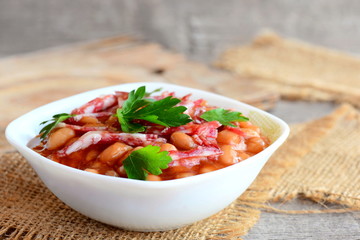 Smoked sausages with white beans in classic tomato sauce. Tasty white beans and smoked sausage stew in a white bowl and on a burlap textile. Rustic style. Home stew