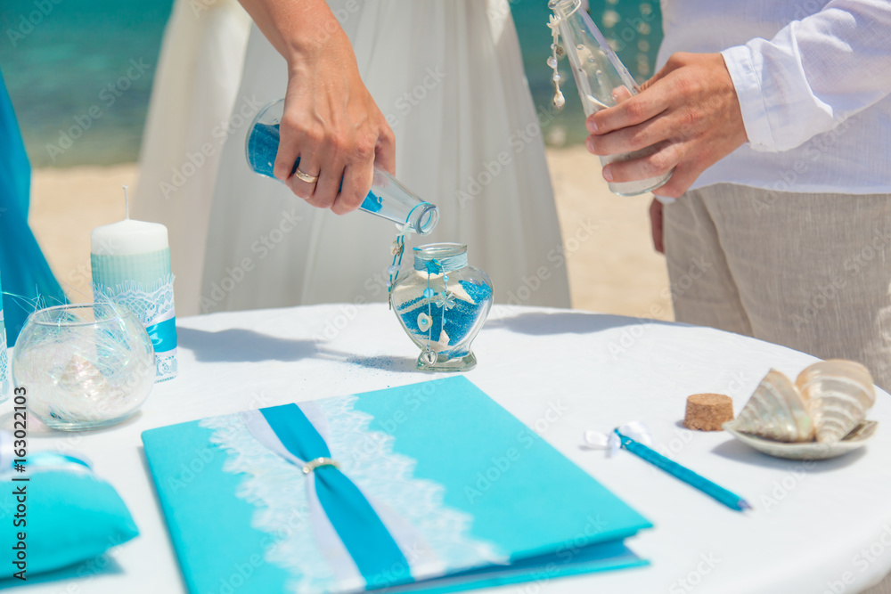 Wall mural Man and woman are pouring blue and white sand into one bottle on the seashore at summer sunny day