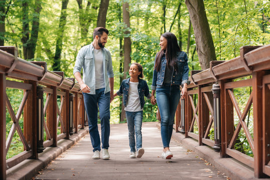 Young smiling interracial family holding hands and walking through the wooden bridge in forest