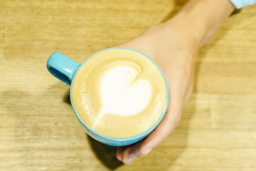 Woman hand holding a blue cup of cappuccino on wooden table background. Heart milk froth.