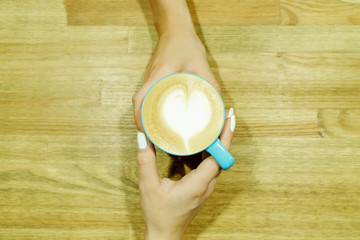 Cappuccino with heart froth between two hands on wooden table. Human pastime