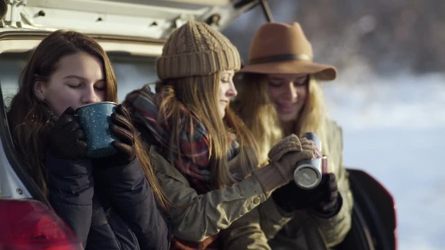 Group Of Teen Girls Hang Out In Back Of Car On Sunny Winter Day, Girl In Middle Pours Her Friends Hot Cocoa, They Cheers And Drink Up