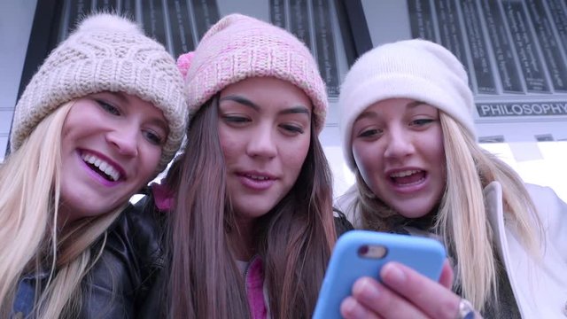 Closeup Of Teens Looking At Something Funny On A Smartphone, They Laugh And Point At The Screen, While Waiting At A Station 