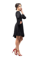 Fototapeta premium Side view of tanned Hispanic woman in black dress posing at camera touching hair. Full body length portrait isolated on white studio background. 