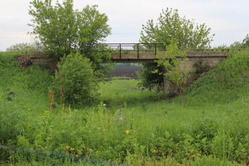 A small railway bridge against a background of green grass and an agricultural field visible in the distance.