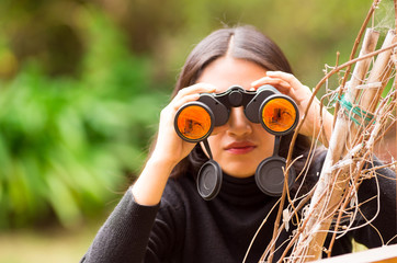 Young woman looking through black binoculars in the forest in a blurred background