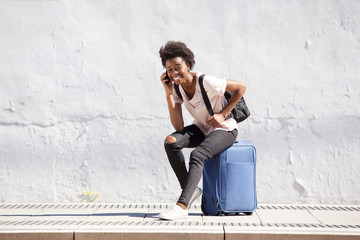 young black woman sitting on suitcase and talking on mobile phone outdoors