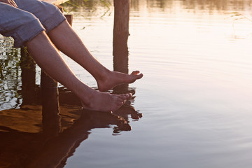 a man sits  on a wooden bridge and dip feet in warm water for a lake in the summer
