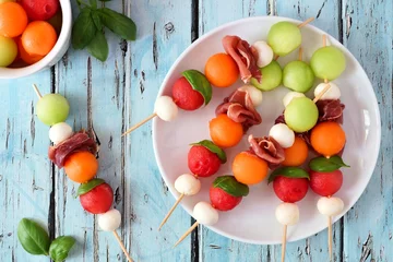 Gartenposter Plate of delicious summer fruit skewers with melon, cheese and prosciutto on a rustic blue wood background © Jenifoto