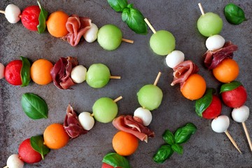 Colorful summer fruit skewers with melon, cheese and prosciutto, overhead view on a rustic metallic tray