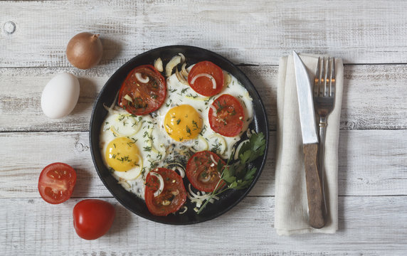 Fried eggs with vegetables and greens (tomatoes, onion, dill) on wooden background with Cutlery on a napkin