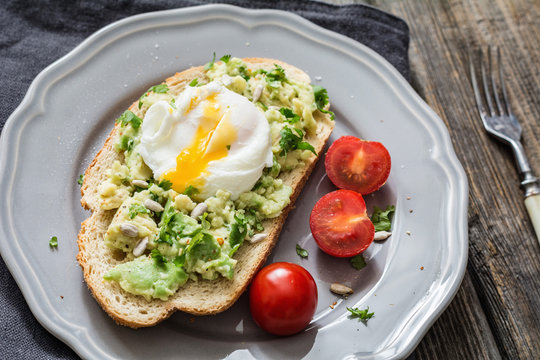 Poached egg on avocado toast. Delicious and healthy snack: mashed avocado with cilantro and sunflower seeds on toasted sourdough bread topped with poached egg on a gray plate. Closeup view