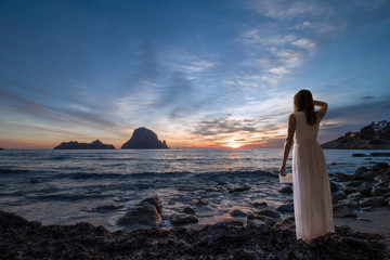 Woman back to the front, dressed in white, watching a sunset and holding a letter