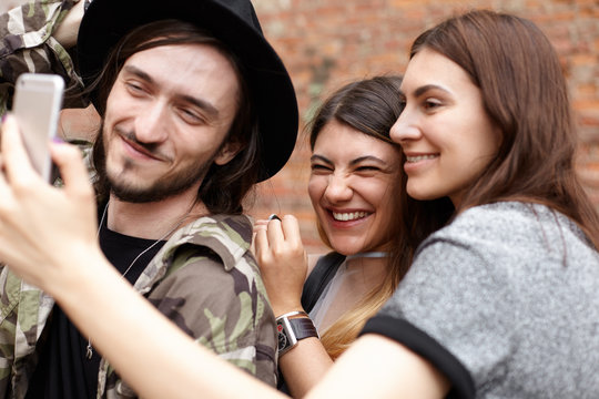 Group of three cheerful best friends laughing, having fun outdoors, taking selfie on mobile phone, sharing pictures via social networks, standing against brick wall background. People and technology