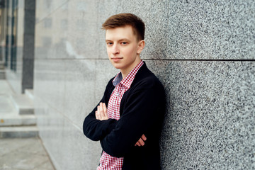 Handsome casual young man in plaid shirt and jacket posing near stone wall outdoors, free space. Teenager with crossed hands. Full body length portrait, fashion and people concept