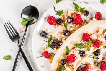Summer fruit berry breakfast. Healthy banana split breakfast with cream cheese, raspberries, blackberries, mint, white and pink chocolate. On white marble plate, marble table. Top view copy space