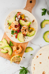 Seafood. Mexican food. Tortilla tacos with traditional homemade salsa salad, parsley, fresh lemon, avocado and grilled shrimp pawns. On a white marble background. Top view copy space