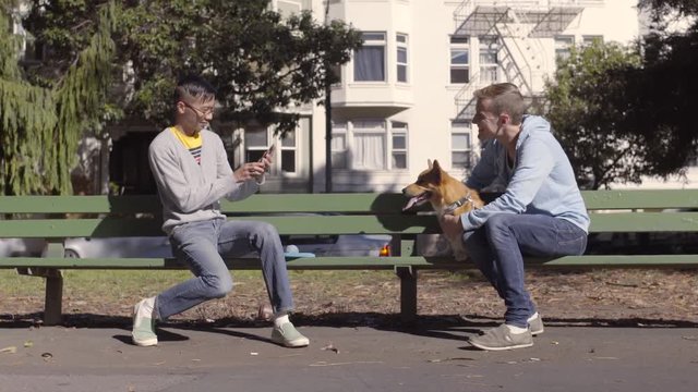 Man Takes Photo Of His Boyfriend And Their Corgi Dog On A Park Bench, Bicyclists Pass By In Foreground