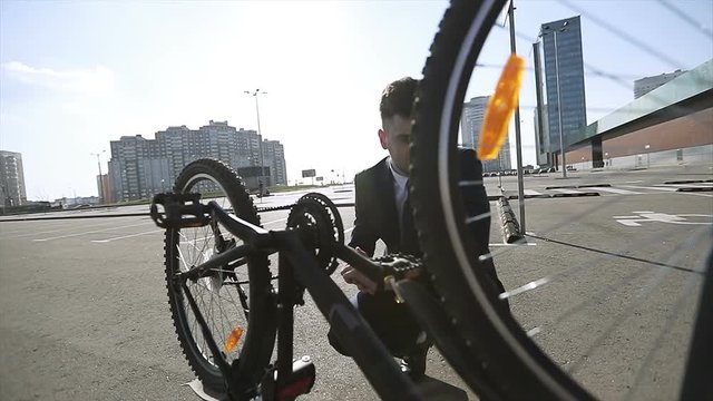 A young businessman in a suit examines an inverted bicycle in sunny weather.