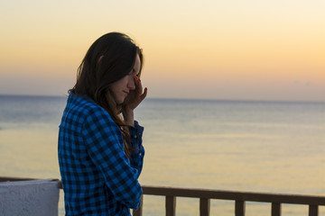 Sleepy young woman standing on balcony by the sea and waiting for the sunrise